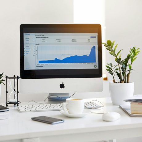 Apple desktop computer displaying a data analysis chart. The computer is standing on a white table surrounded with assorted white color inspired decor